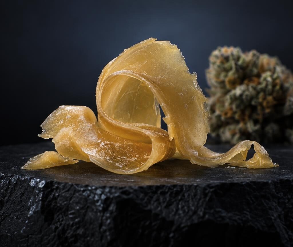 Golden translucent Rosin shards on dark stone surface, cannabis flower in background